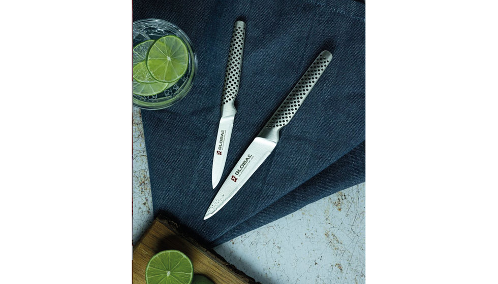Two stainless steel pairing knives from Global on a tablecloth next to a Gin and Tonic in a small tumbler with a slice of lime. 