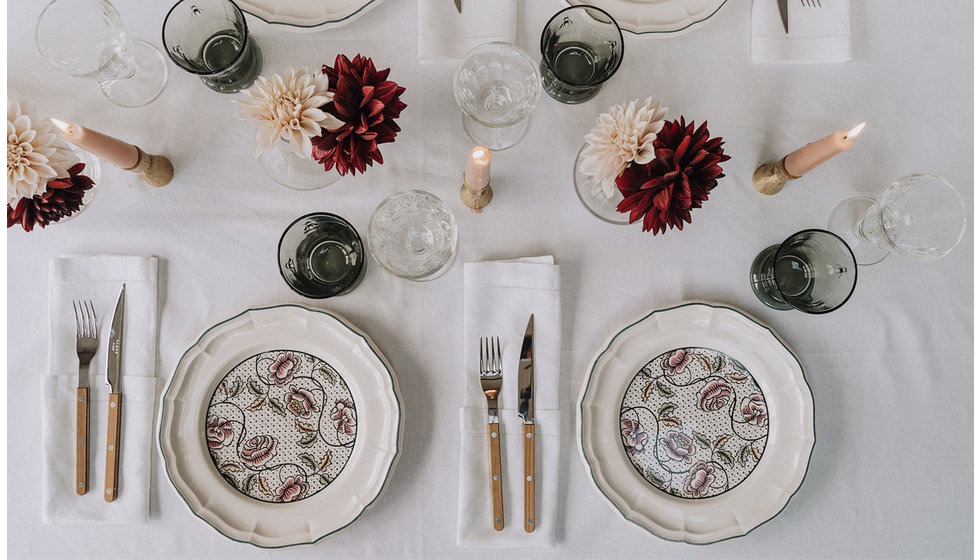 A chic dinner table laid with a white linen tablecloth, Gien dinner plates, green water tumblers and peach and burgundy dahlias in small bud vases. 