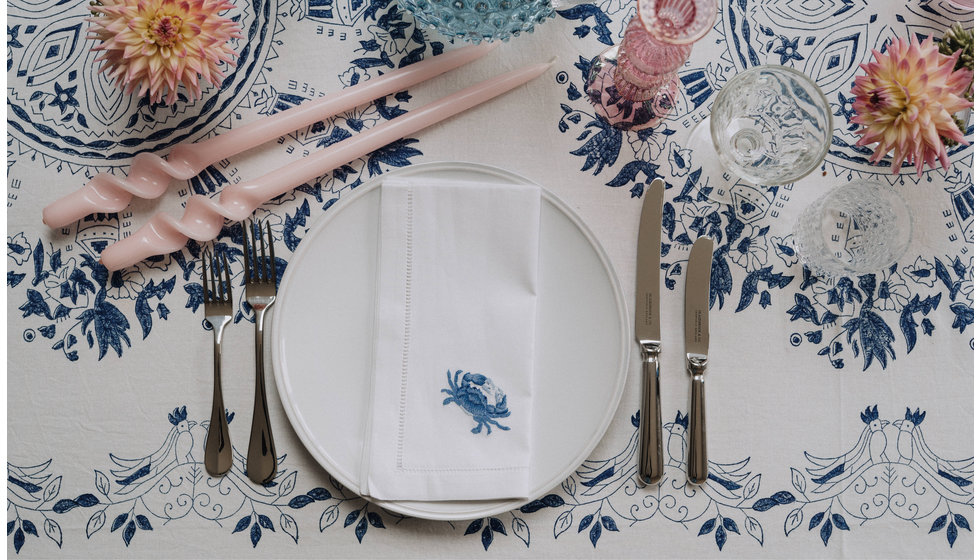 A colourful tablescape with a Penny Morrison tablecloth, plain Costa Nova dinner plates, silver cutlery and an embroidered white linen napkin with a shell on it stitched in blue. 