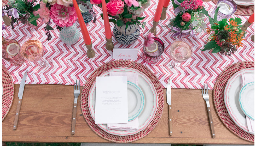 A colourful al fresco table laid with a Wicklewood printed tablecloth, pink rattan placemats, Gien scalloped dinner plates, wooden cutlery from Sabre and colourful cut glass from Gurasu. 