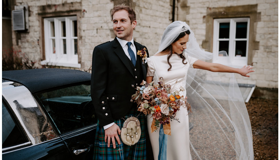 Sophie and Graeme stand next to their wedding car - a Bentley. Sophie adjusts her veil.