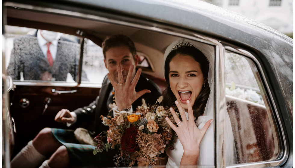 Sophie and Graeme sat in the back of a car both playfully holding up their handing with their new wedding rings on and smling. 