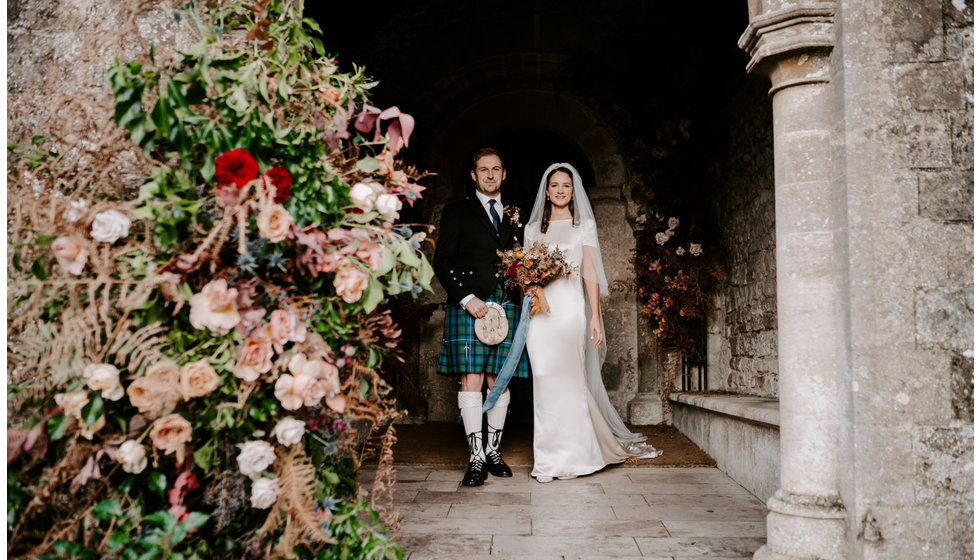 Sophie and Graeme next to a beautiful arch of winter flowers on the outside of the Church.