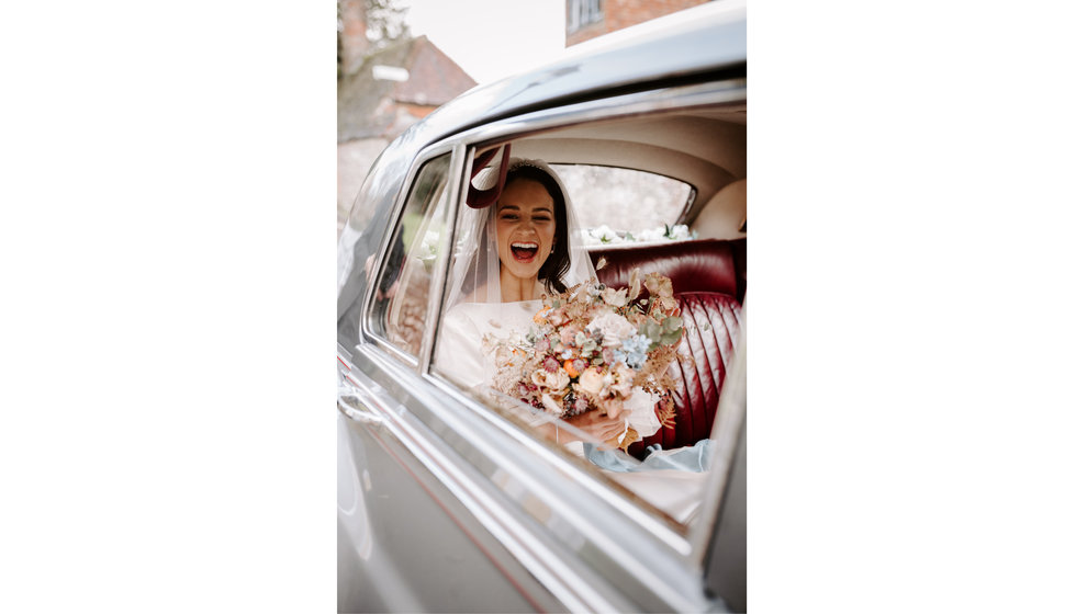 Bride Sophie sits in the back of a Bentley on the way to the Church. She holds her bouquet of wintery florals and smiles.