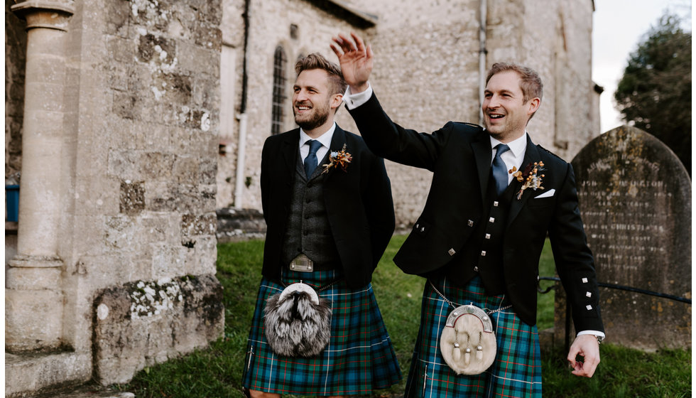 The groom and his best man stand outside the church wearing traditional tartan kilts.