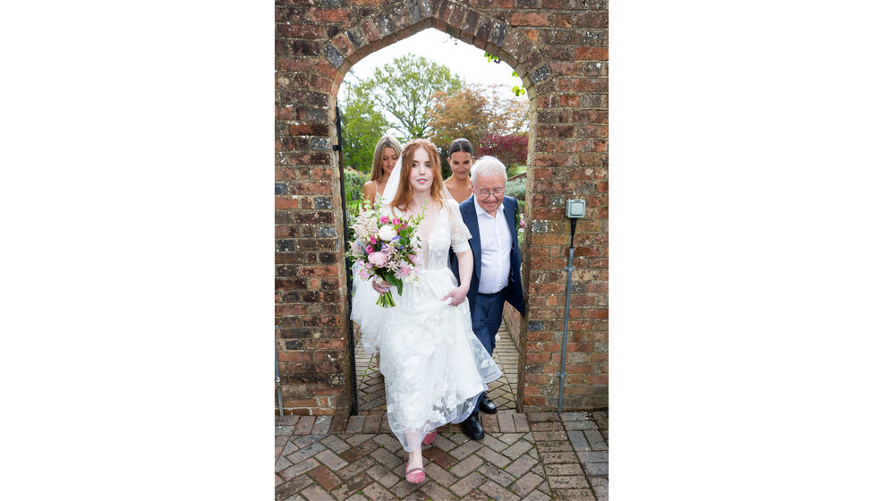 Bride sarah holds her wedding dress and walks through an exterior arch with her father.
