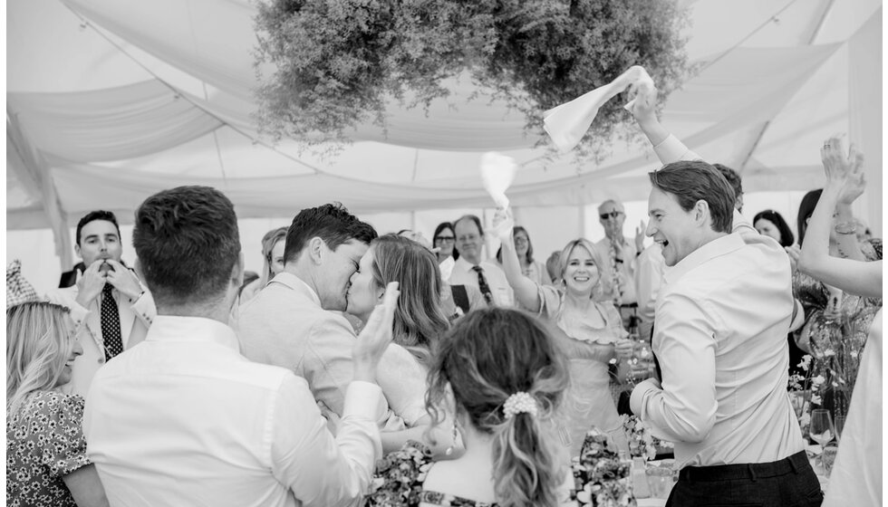 Charlotte & Harry's Magical Meadow Marquee Wedding in Oxfordshire: Bride and groom kissing while their wedding guests are waving white napkins