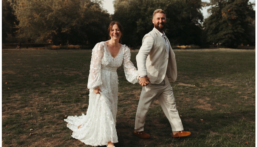 Whimsical Floral Wedding in London Park | The Smiling bride in a floral dress and the groom in a linen suit are holding hands together in Myatt's Field Park.