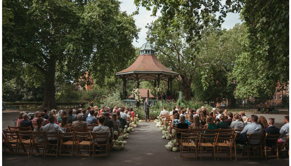 Whimsical Floral Wedding in London Park | Wedding ceremony with officiant and wedding guests in front of a park bandstand decorated with white flowers