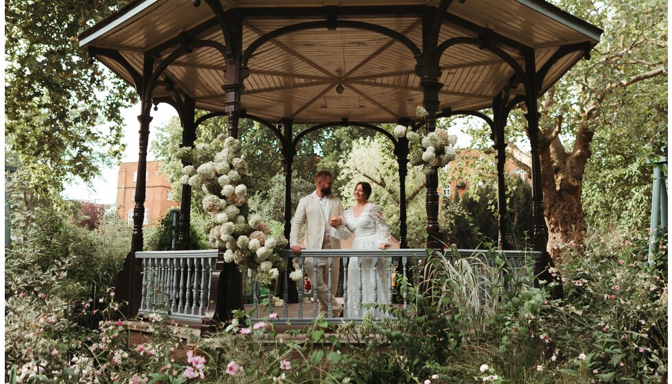 Whimsical Floral Wedding in London Park | The bride in a floral white dress and the groom in a cream linen suit are standing on a park bandstand that is decorated with seasonal white flowers and lots of green foliage.