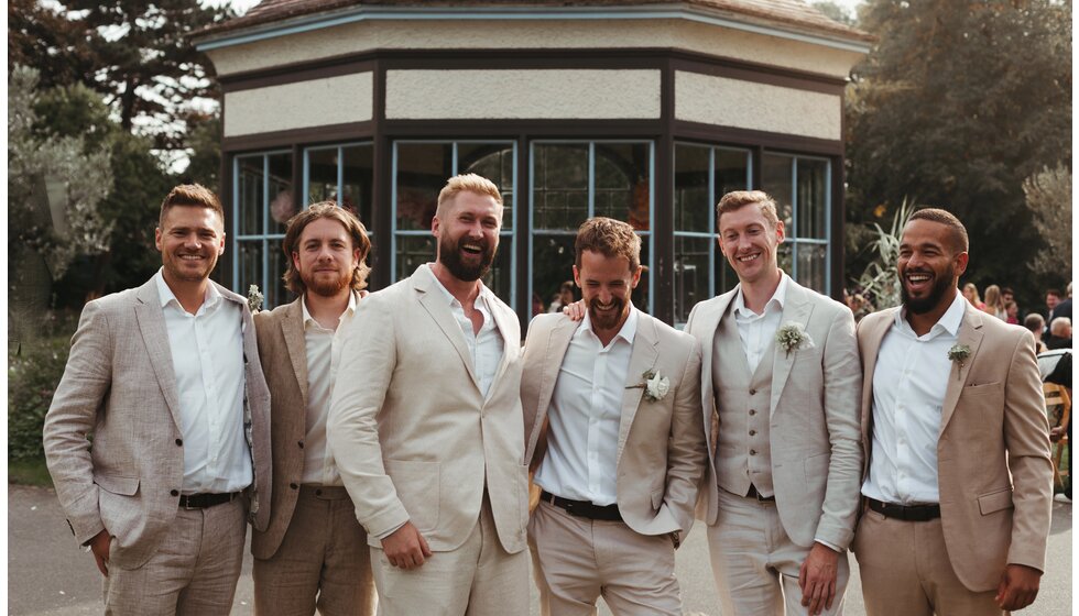 Whimsical Floral Wedding in London Park | The smiling groom and the groom's party wearing matching beige suits are standing next to each other in Myatt's Field Park.