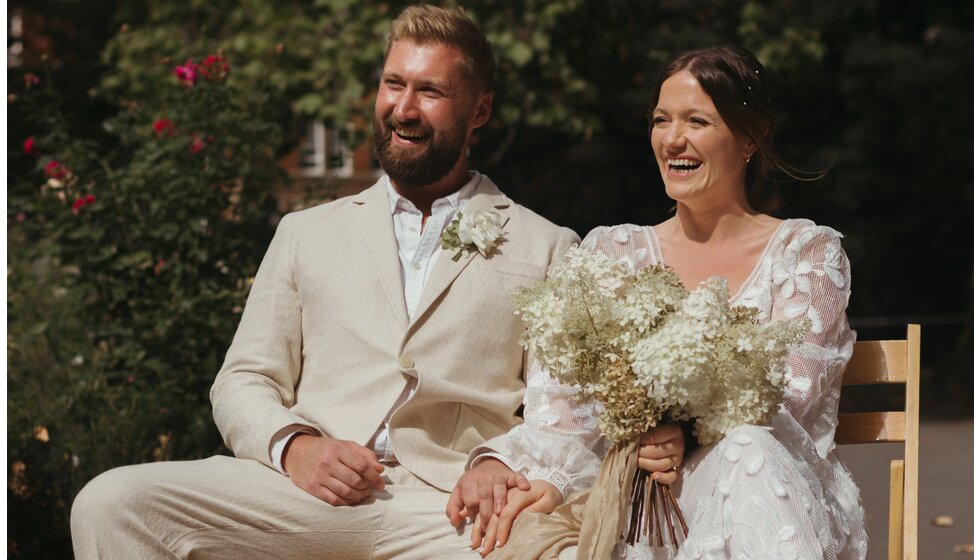 Whimsical Floral Wedding in London Park | The smiling bride in a floral wedding dress and the groom in a linen suit are sitting during a wedding ceremony in Myatt's Field Park, holding hands together.


