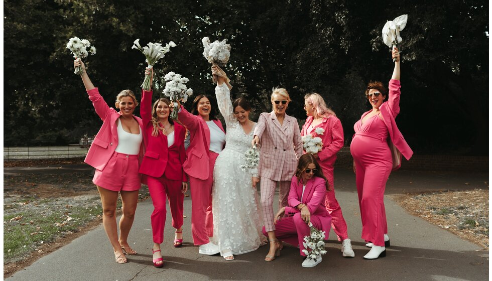 Whimsical Floral Wedding in London Park | The bride posing with her bridal party wearing matching pink bridesmaids suits and holding white flowers in the park