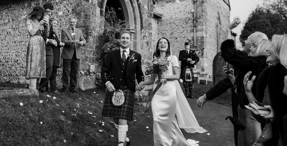 Sophie and Graeme walking out of the Church. Sophie wears a short sleeve silk dress, Graeme wears a traditional kilt in family tartan. The couple hold hands and smile as they walk past their guests.