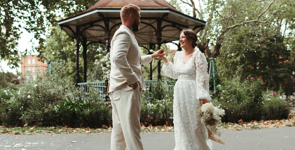 Whimsical Floral Wedding in London Park | The bride in a floral dress and the groom in a linen suit are holding hands in front of a park bandstand.